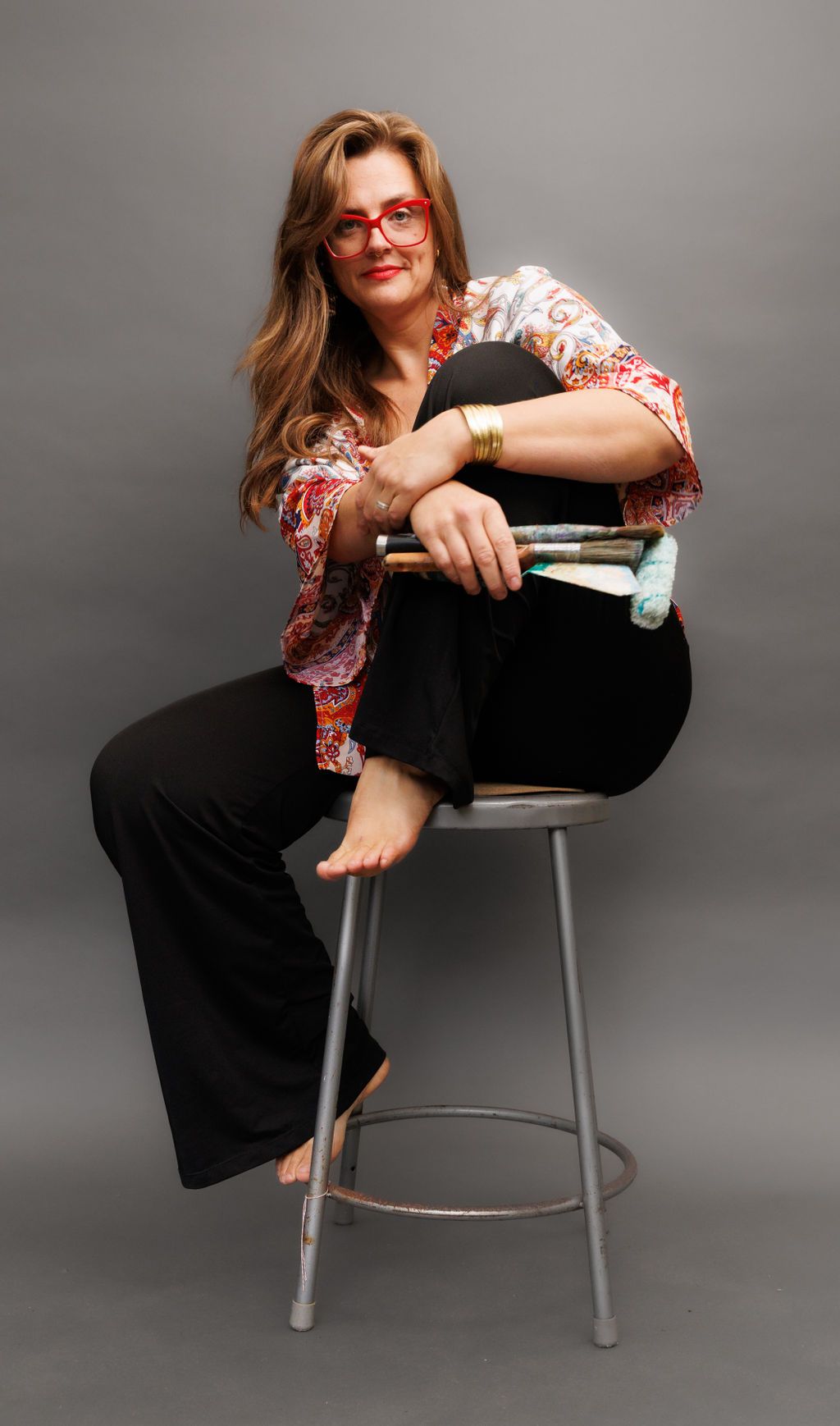 Hope Black, long wavy hair, sitting on a stool barefoot, smiling at the camera, with paint brushes and a roller in one hand. One knee up, in a pose of nonchalance but with a welcoming air. I love you!