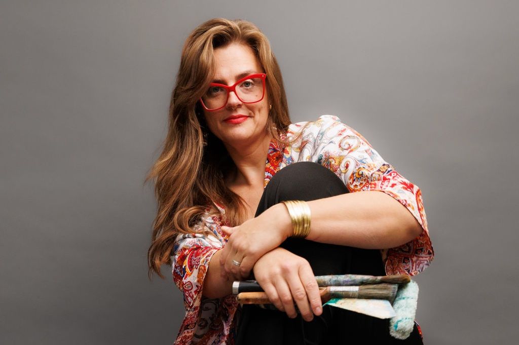 Hope Black, long wavy hair, sitting on a stool barefoot, smiling at the camera, with paint brushes and a roller in one hand. One knee up, in a pose of nonchalance but with a welcoming air. I love you!